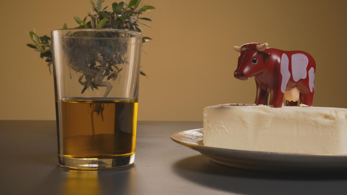 Sur la gauche, un verre d'huile d'olive (posée devant un brin d'olivier) et sur la droite une assiette contenant une motte de beurre sur laquelle trône une vache (un jouet pour enfant type Duplo)